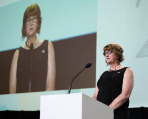 Jodi-Gray-wearing a black dress and speaking in an event