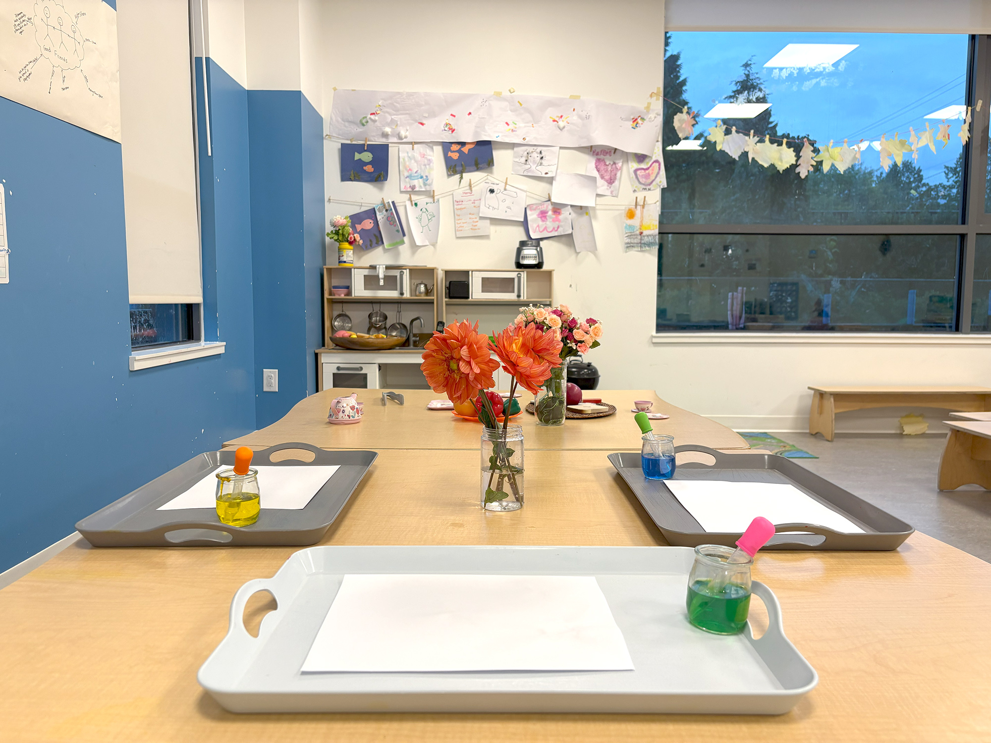 A large wooden table with flowers, white paper, and colored inks for drawing and coloring, with a window overlooking greenery, children’s artwork on the wall, and toys placed along a blue wall