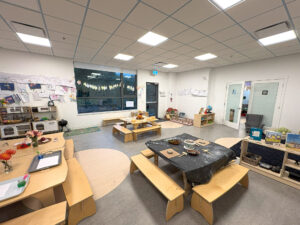 A large children’s play and activity room with white walls, a large window, children’s artwork displayed on the walls, and multiple wooden tables with flowers and toys