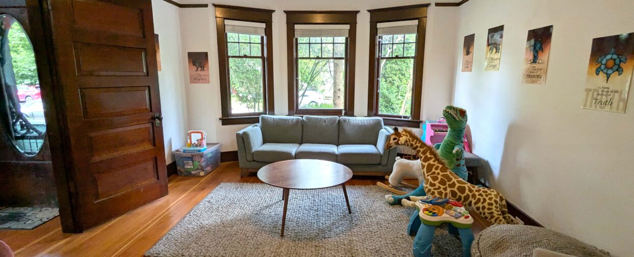 Living room with three windows facing greenery, a gray three-seater sofa, toys placed along the wall, posters on the right wall, and a door on the left side.